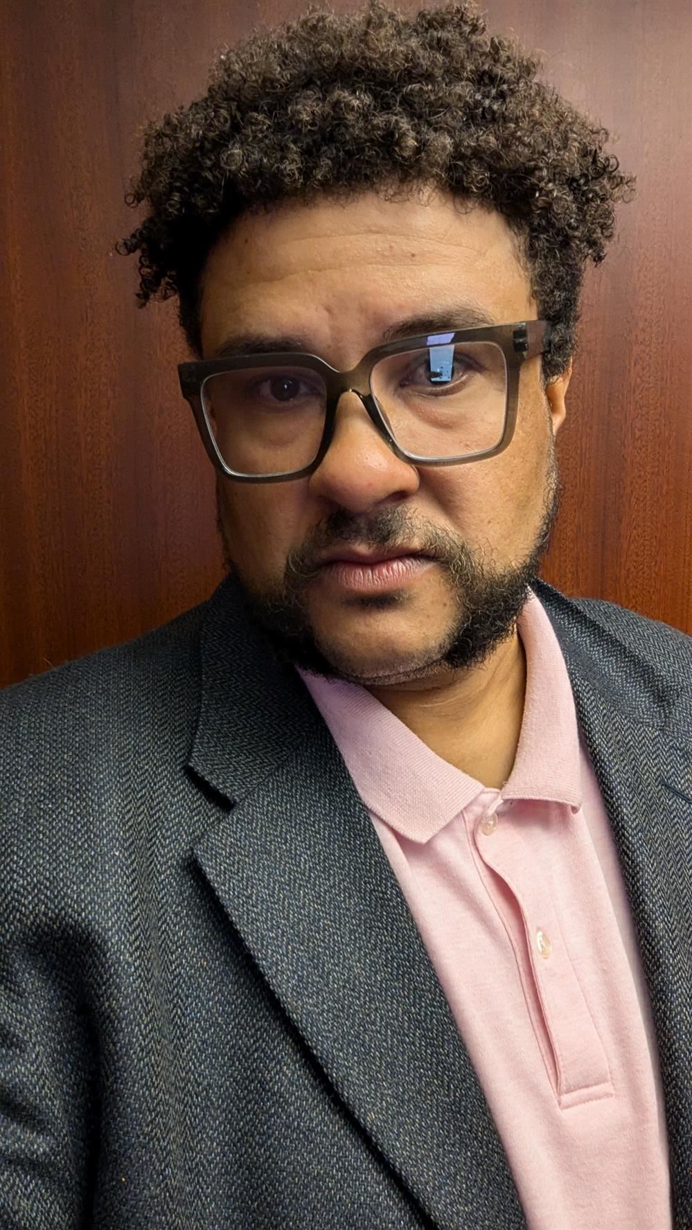 A tall and broad-set man wearing a pink polo shirt and twill-fabric blazer looks into the camera while wearing glasses